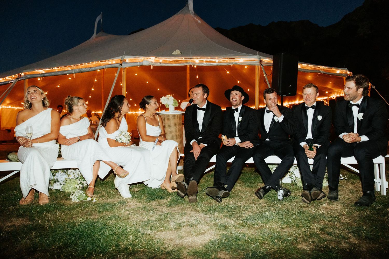 Wedding party poses together under illuminated tent with bridesmaids and groomsmen in formal attire.