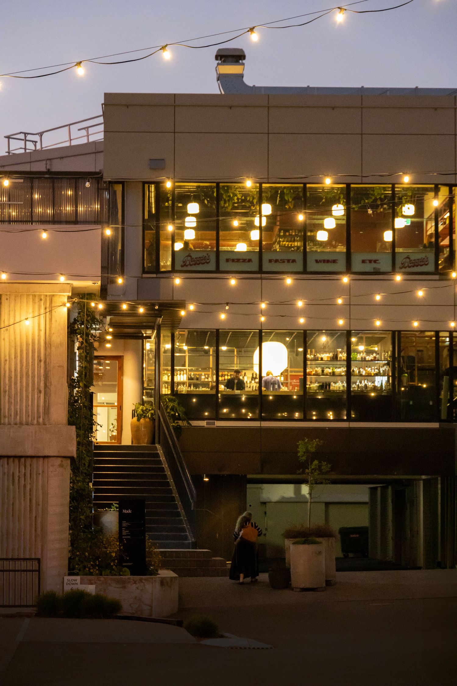 Evening glow on modern glass restaurant with warm hanging lights and upper-level seating.