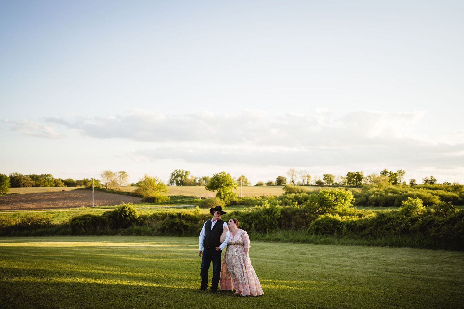 A couple stands together on a lush green hill overlooking a scenic countryside landscape at sunset.