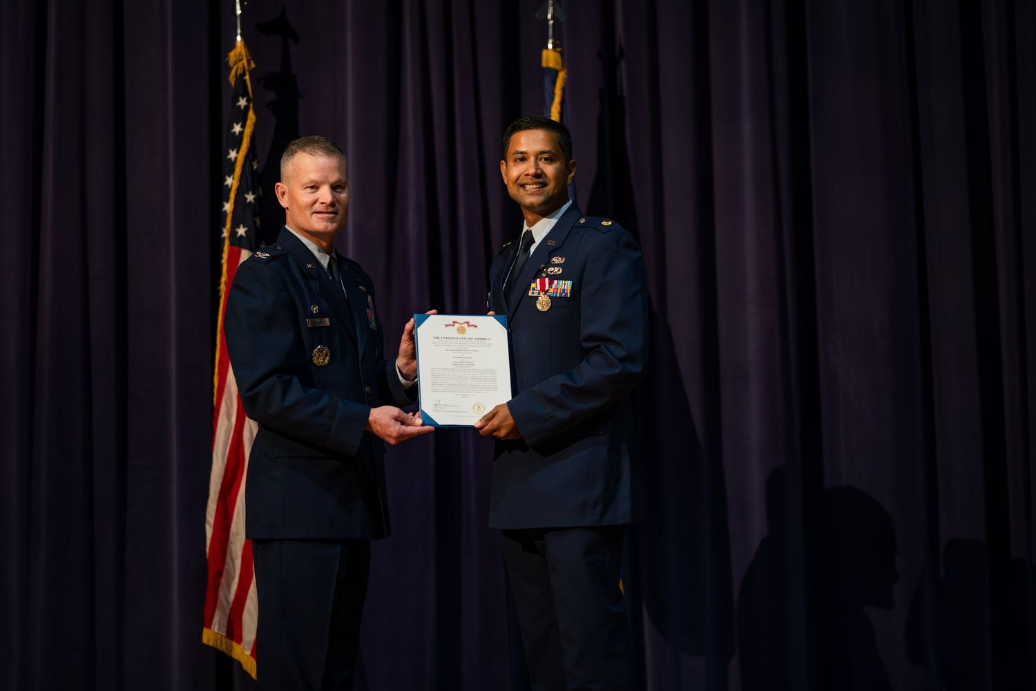 Two military officers in dress uniforms exchange documents during formal ceremony with flags.