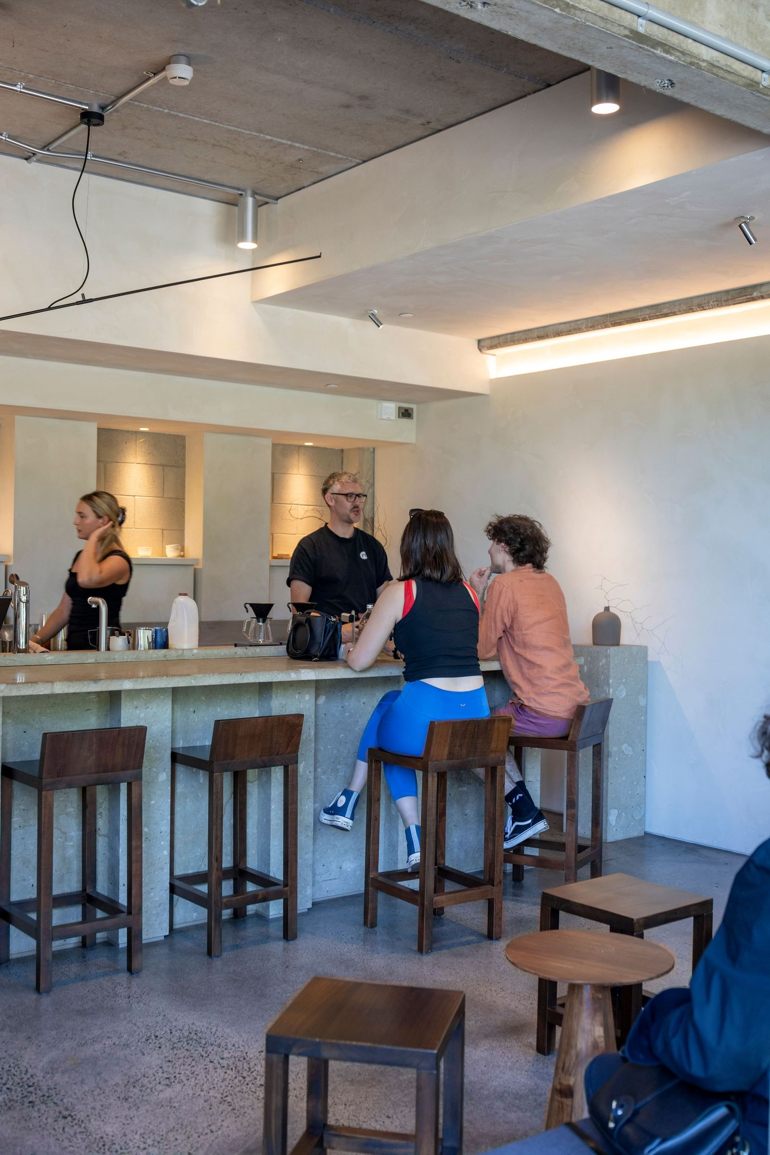 Minimalist café with a concrete bar, stools, and a small group chatting with the barista.