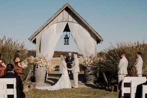 A rustic outdoor wedding ceremony takes place at a charming white chapel surrounded by tall grasses and seated guests.