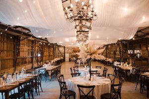 An elegant barn reception venue with rustic chandeliers, draped ceiling, and round tables set for a wedding celebration.