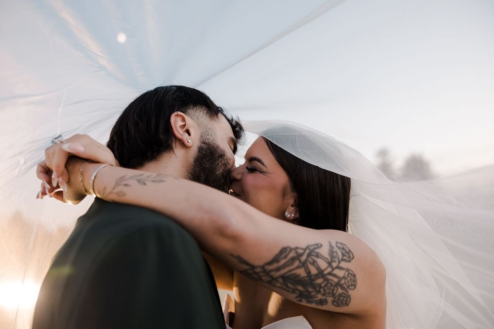 Romantic couple share intimate moment under flowing veil during outdoor sunset wedding celebration.