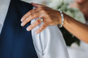 Close up detail of a hand wearing an engagement ring and delicate bracelet against a navy blue suit.