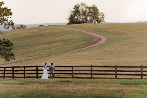 Bride and groom stand by a wooden fence on a scenic farm with a winding dirt path and rolling green hills.