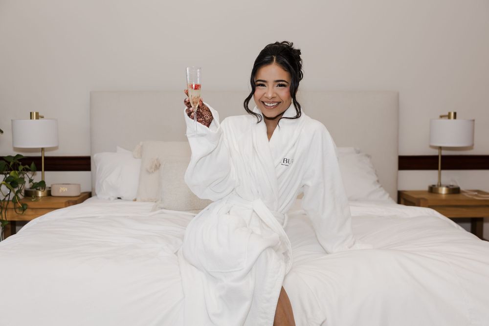 Smiling bride in white robe holding champagne glass while sitting on hotel bed with white bedding.