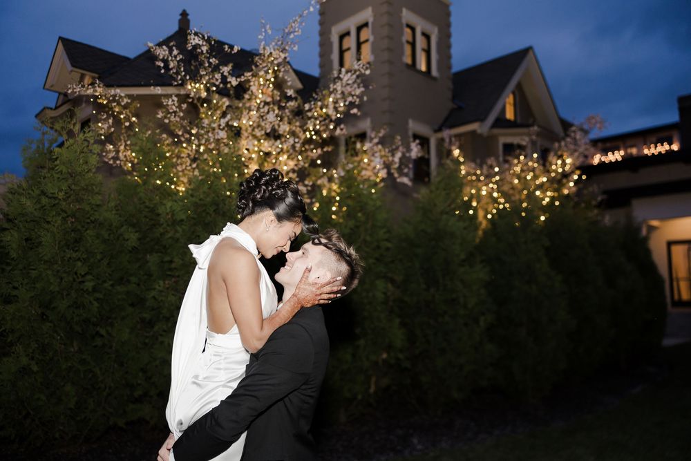 Newlywed couple sharing a romantic kiss outdoors at night with a decorated Victorian mansion in the background.