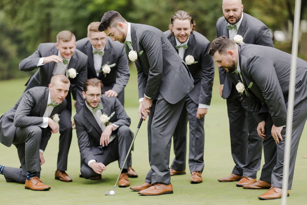 Groomsmen in gray suits and brown shoes crouched down in a fun lineup pose outdoors on grass.