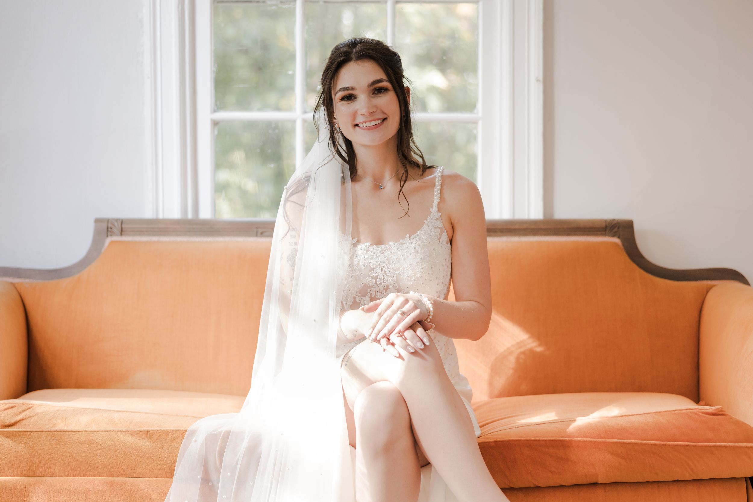 Bride in white lace gown and veil sits smiling on an orange velvet sofa in a bright, elegant room.