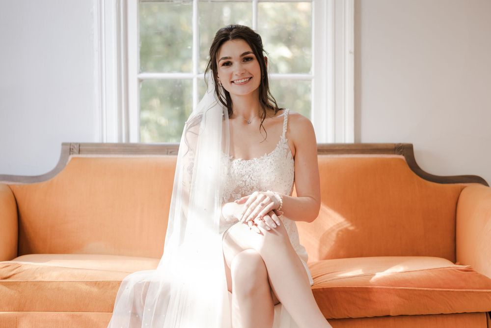 Bride in white lace gown and veil sits smiling on an orange velvet sofa in a bright, elegant room.