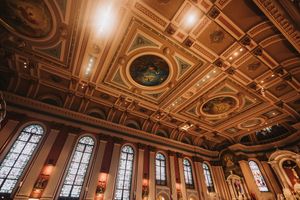 Ornate gilded ballroom ceiling with circular medallions, chandeliers and tall arched windows.
