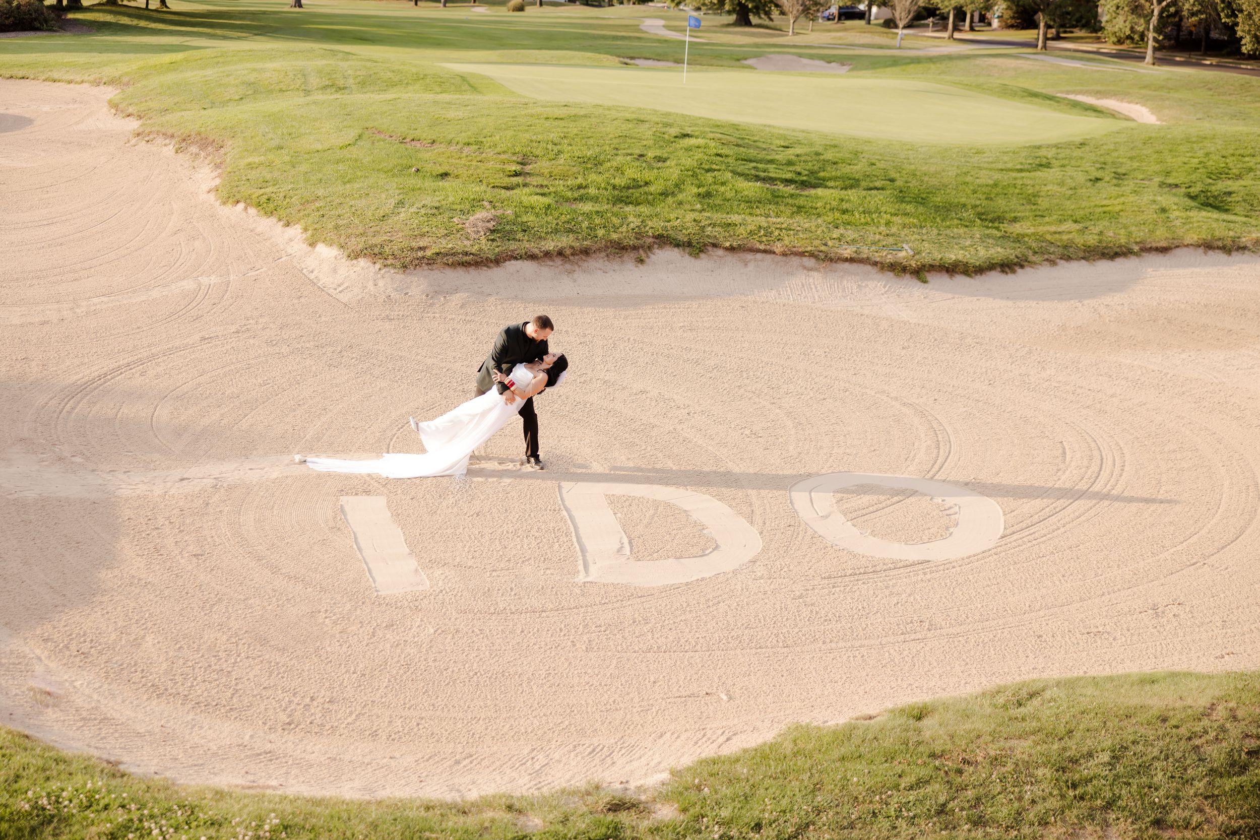 Golfer takes a swing from a sand bunker on a scenic golf course surrounded by green grass and trees.