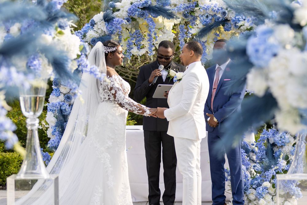An elegant outdoor wedding ceremony setup with blue floral decor and white chairs under a sunny sky.