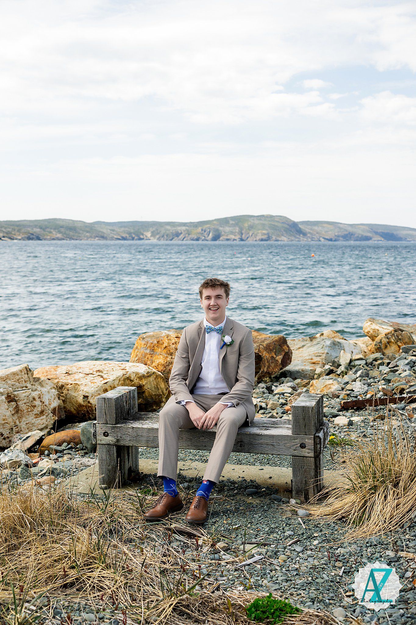Someone in a light gray suit sits on a wooden bench along a rocky coastline with blue water and hills in background.