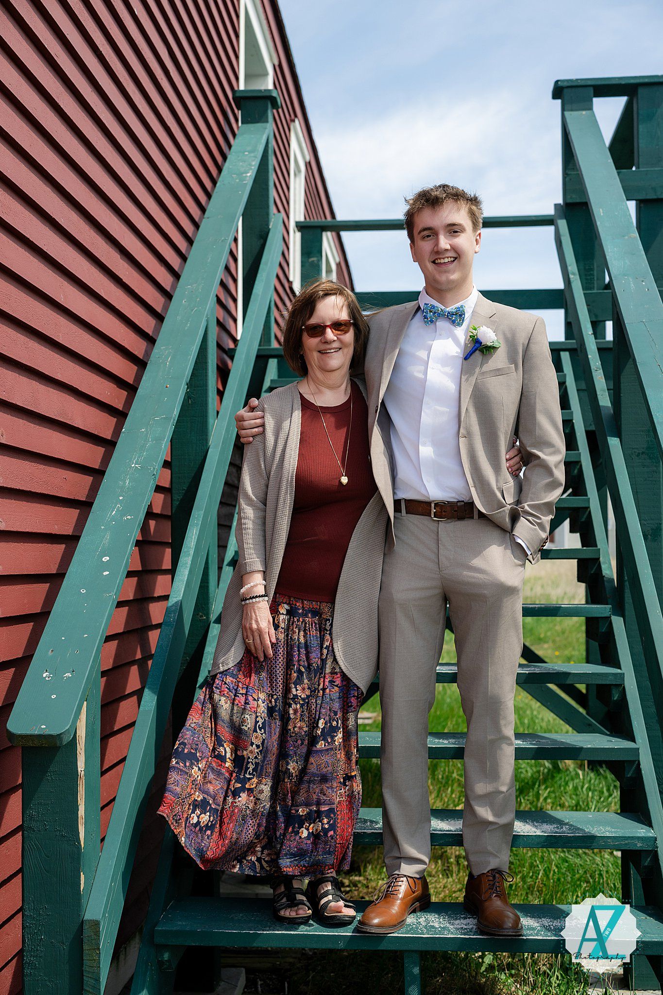 Two people stand on green wooden stairs against a red barn wall wearing formal attire with a blue bow tie and floral skirt.