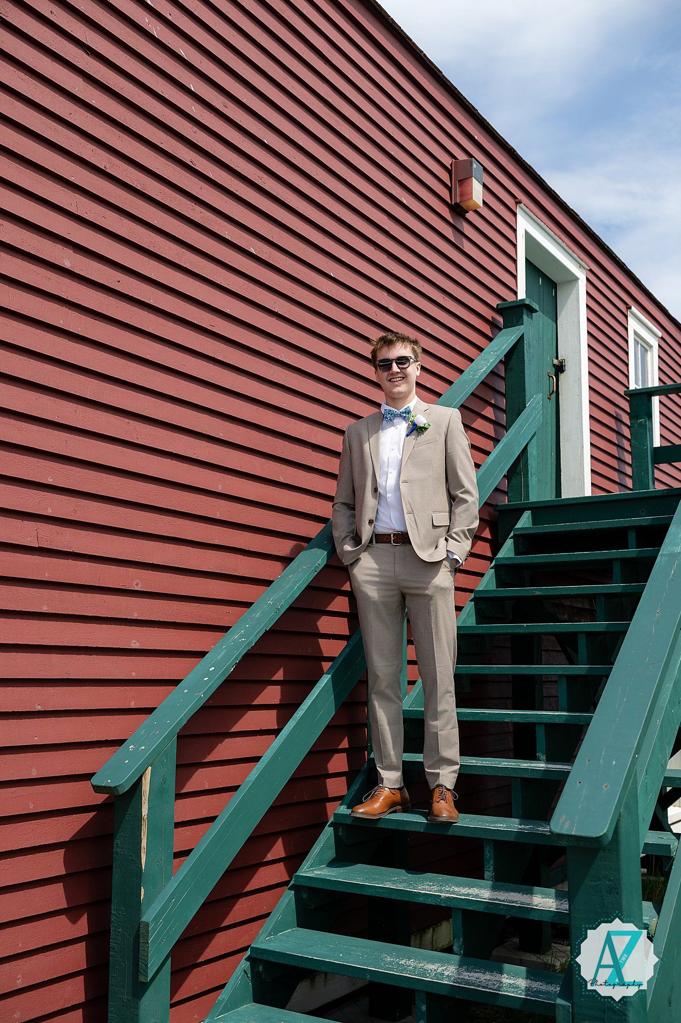 A person in a light suit and bowtie poses on green wooden stairs against a red barn-style building exterior.