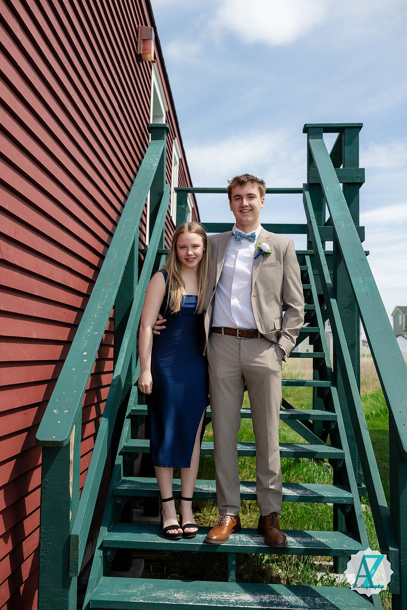 Two people stand together on green metal stairs against a red barn wall under a blue sky on a sunny day.