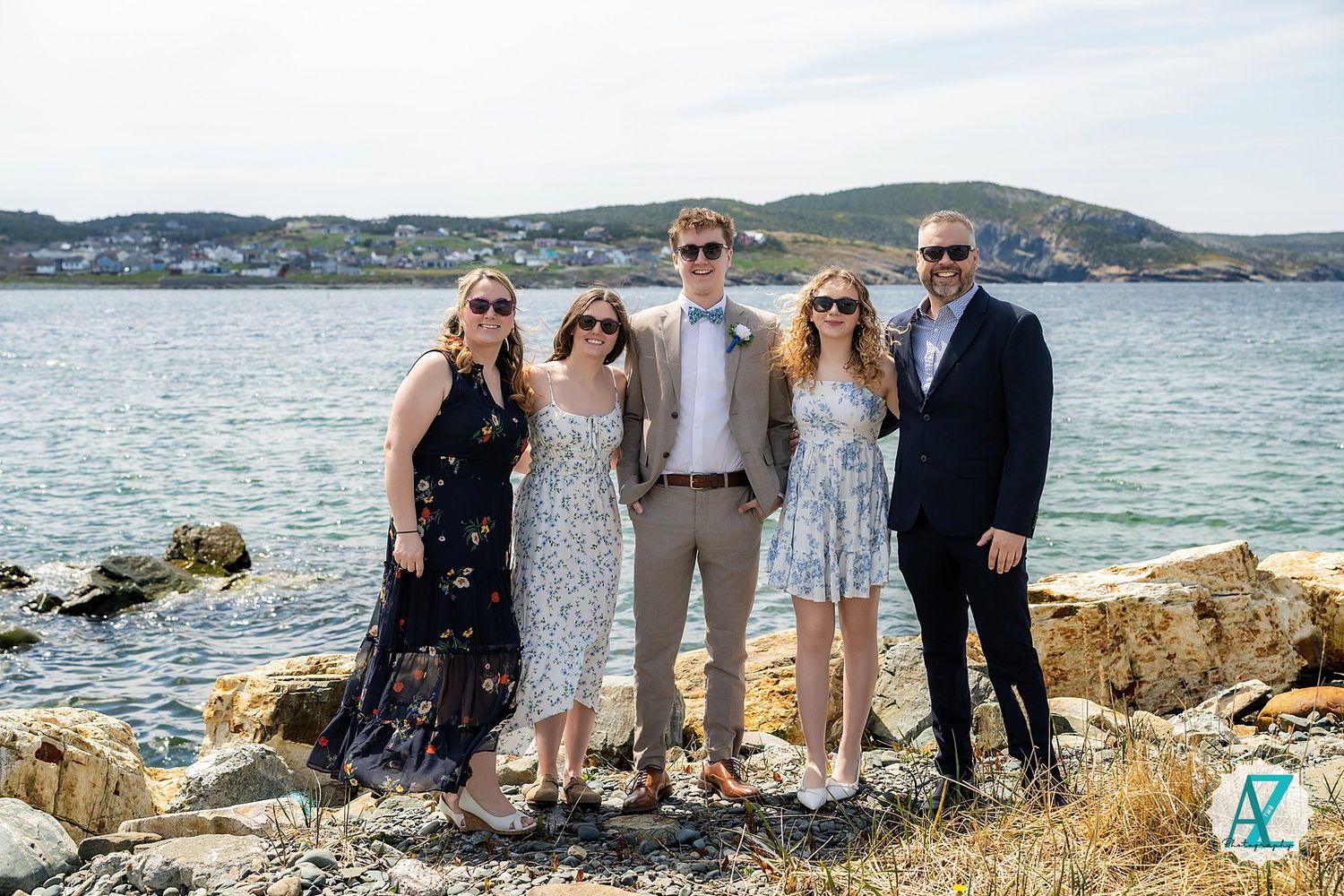 A group of five people in formal attire pose together on a rocky coastline with blue water and mountains in the background.