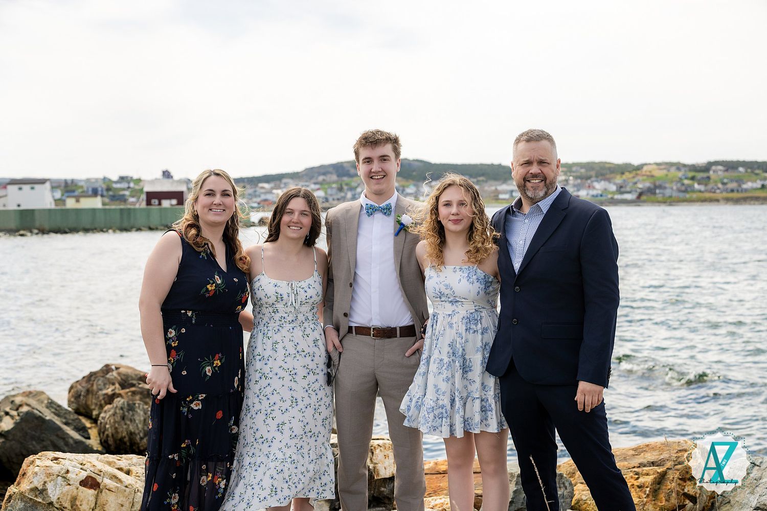 Five people in formal attire stand together on a rocky coast with the ocean and coastal town in the background.
