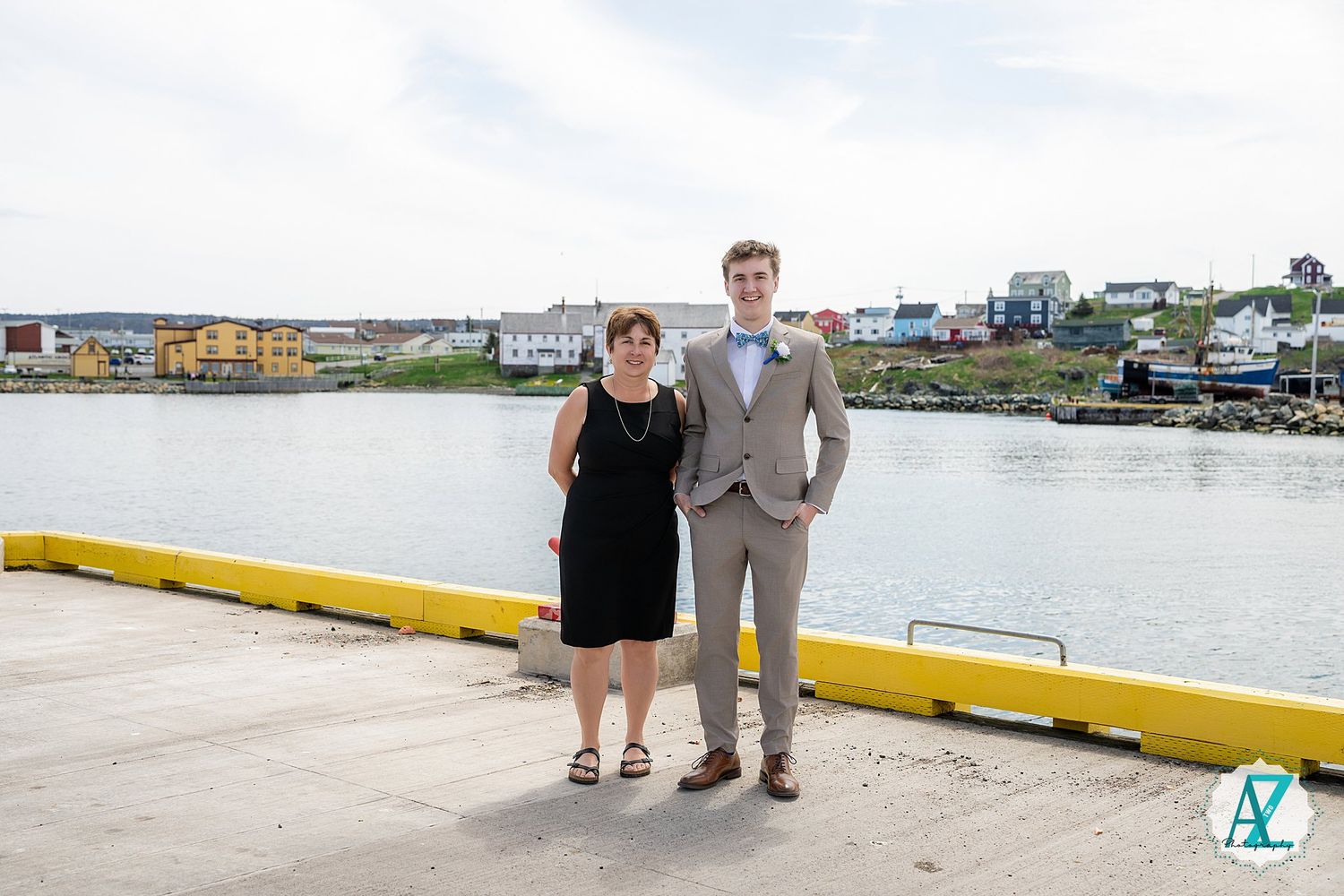 A couple stands on a pier overlooking a waterfront with colorful buildings in the background on a bright day.