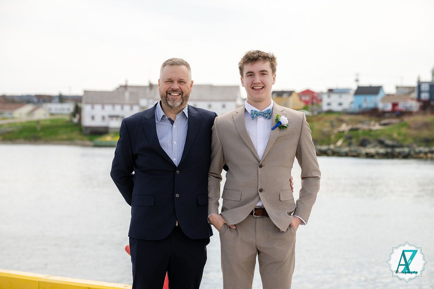 Two people in formal attire stand together smiling by the waterfront on a sunny day.
