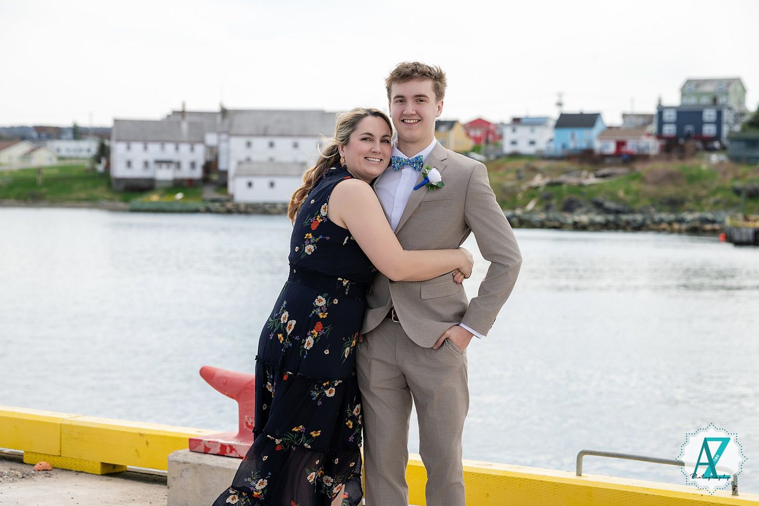 A couple in coordinating formal outfits poses by a waterfront dock area in overcast weather.