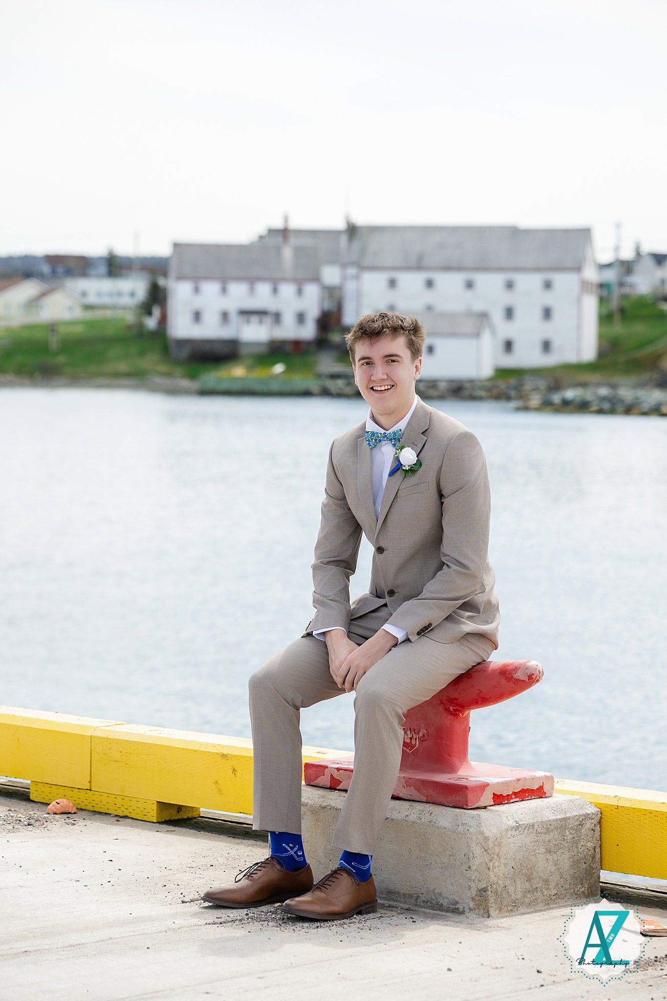A person in a tan suit and bow tie sits on a red bollard by the waterfront with white buildings in the background.