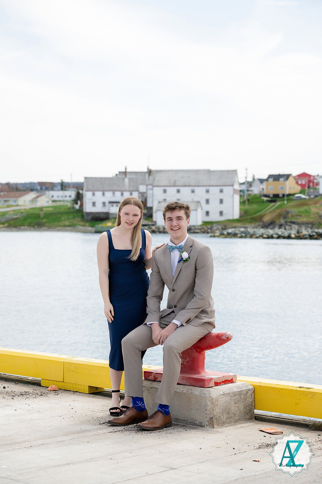 Couple in formal attire pose by waterfront with white buildings and red boat dock in Newfoundland background.