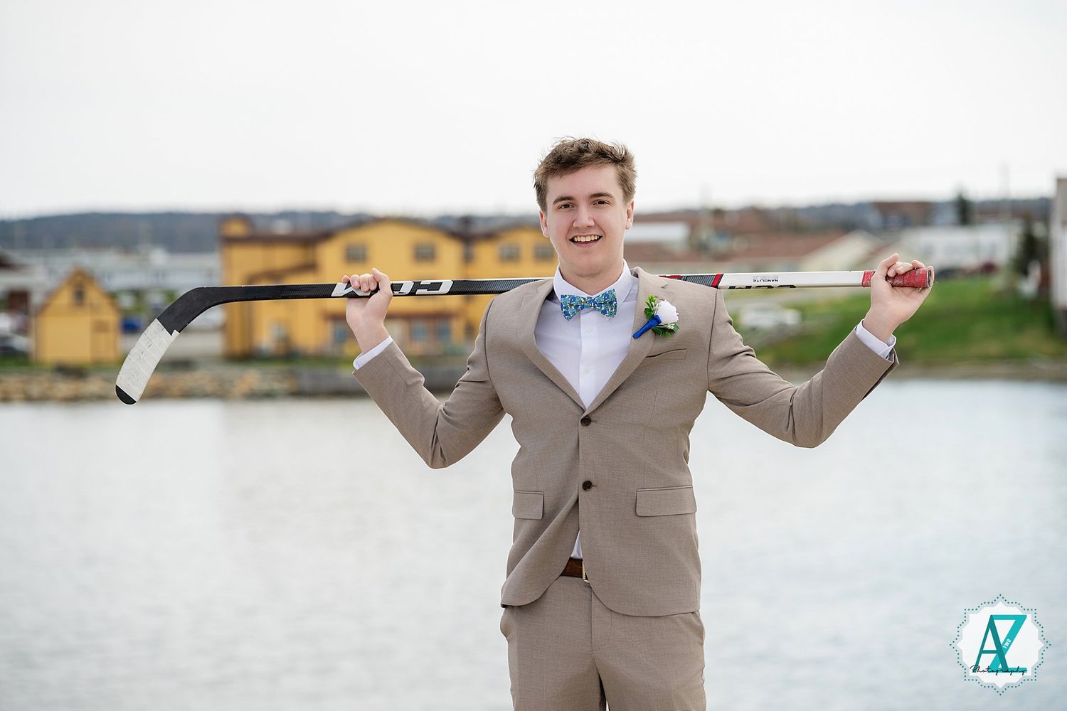 A person in a beige suit and blue bowtie poses playfully with a fishing rod by the water.
