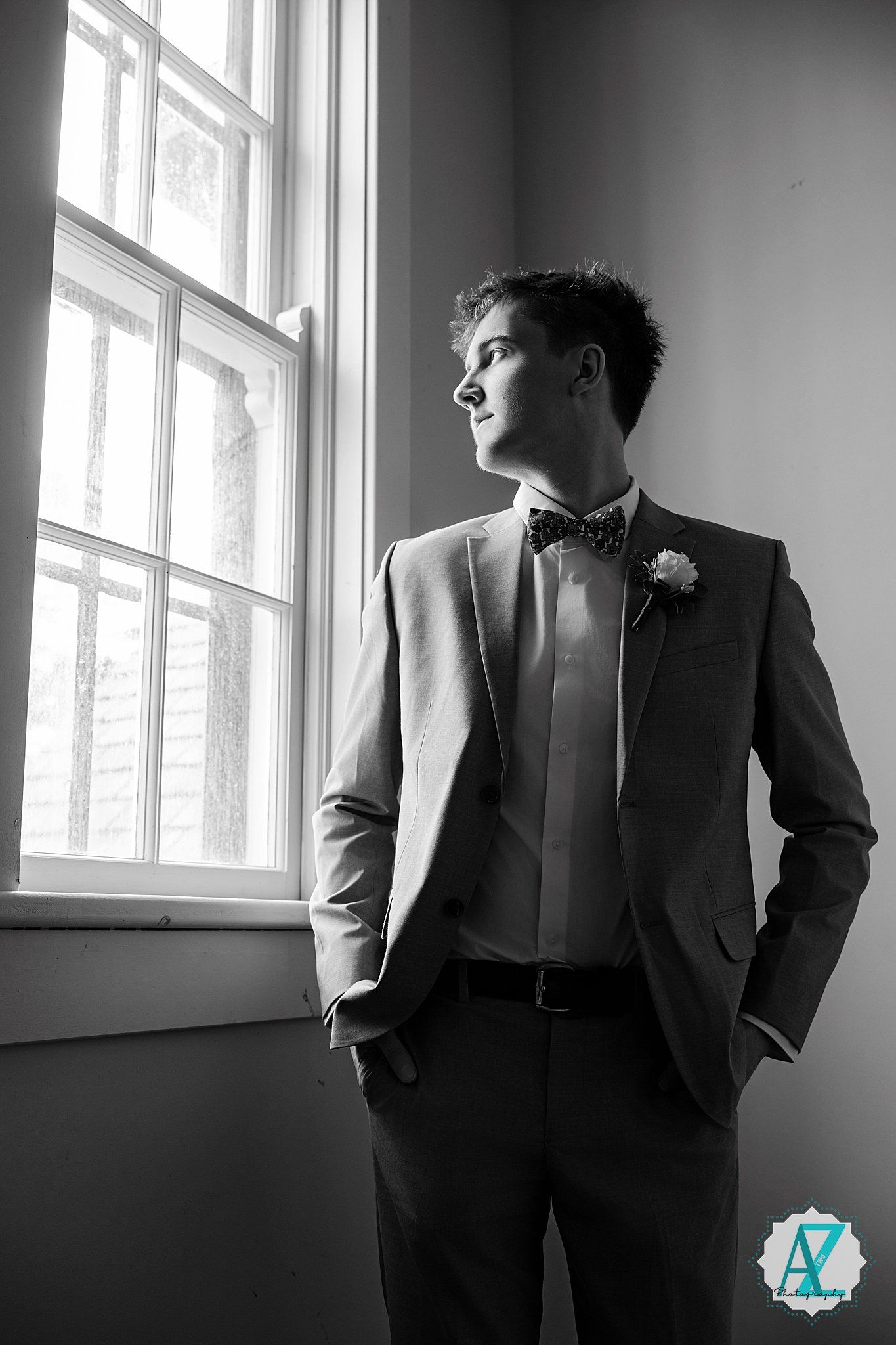 A person in formal attire with a bowtie and boutonniere gazes thoughtfully out of a bright window in black and white.