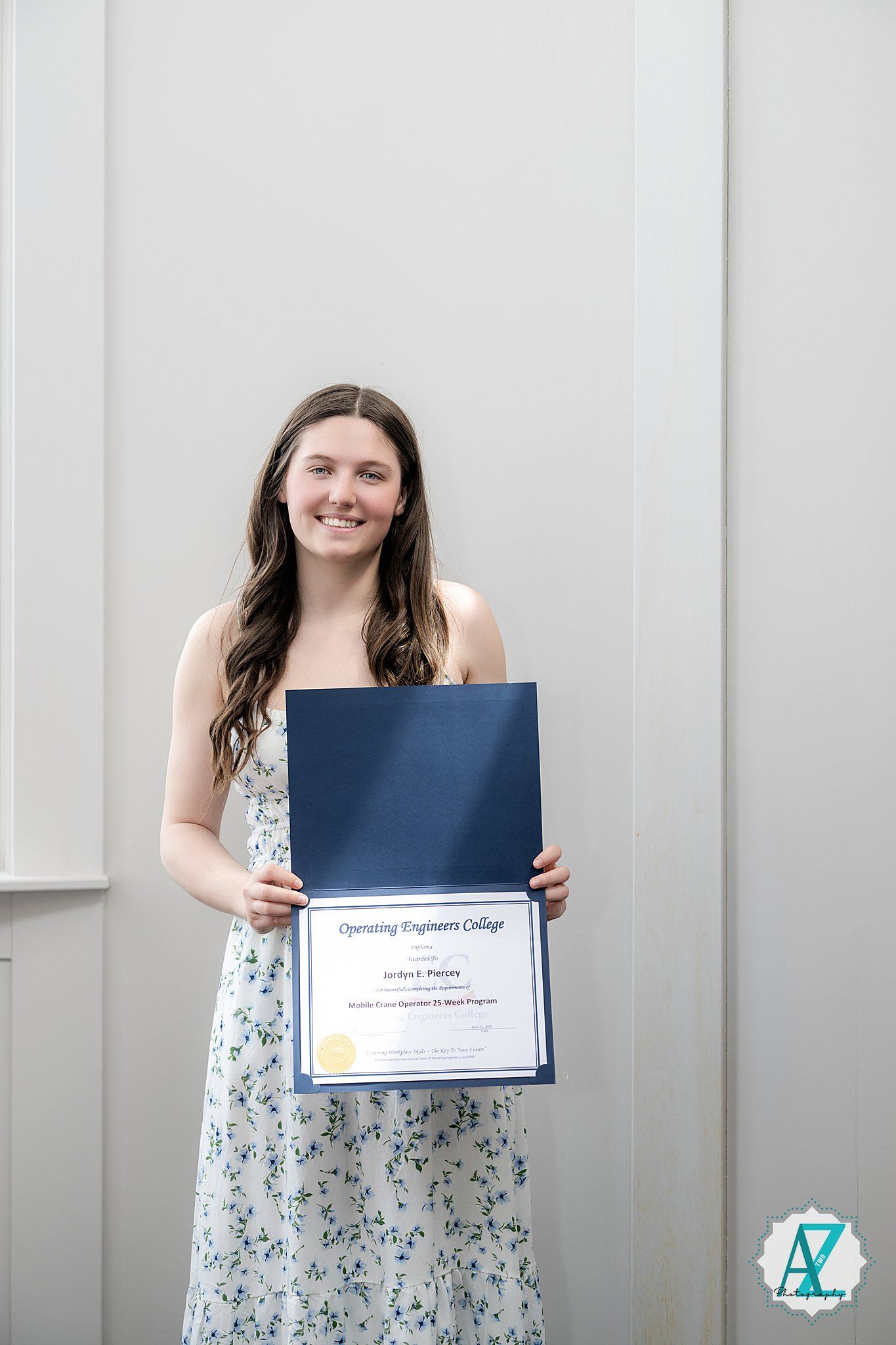 A person in a floral dress holds a blue diploma or certificate while standing against a light gray wall.