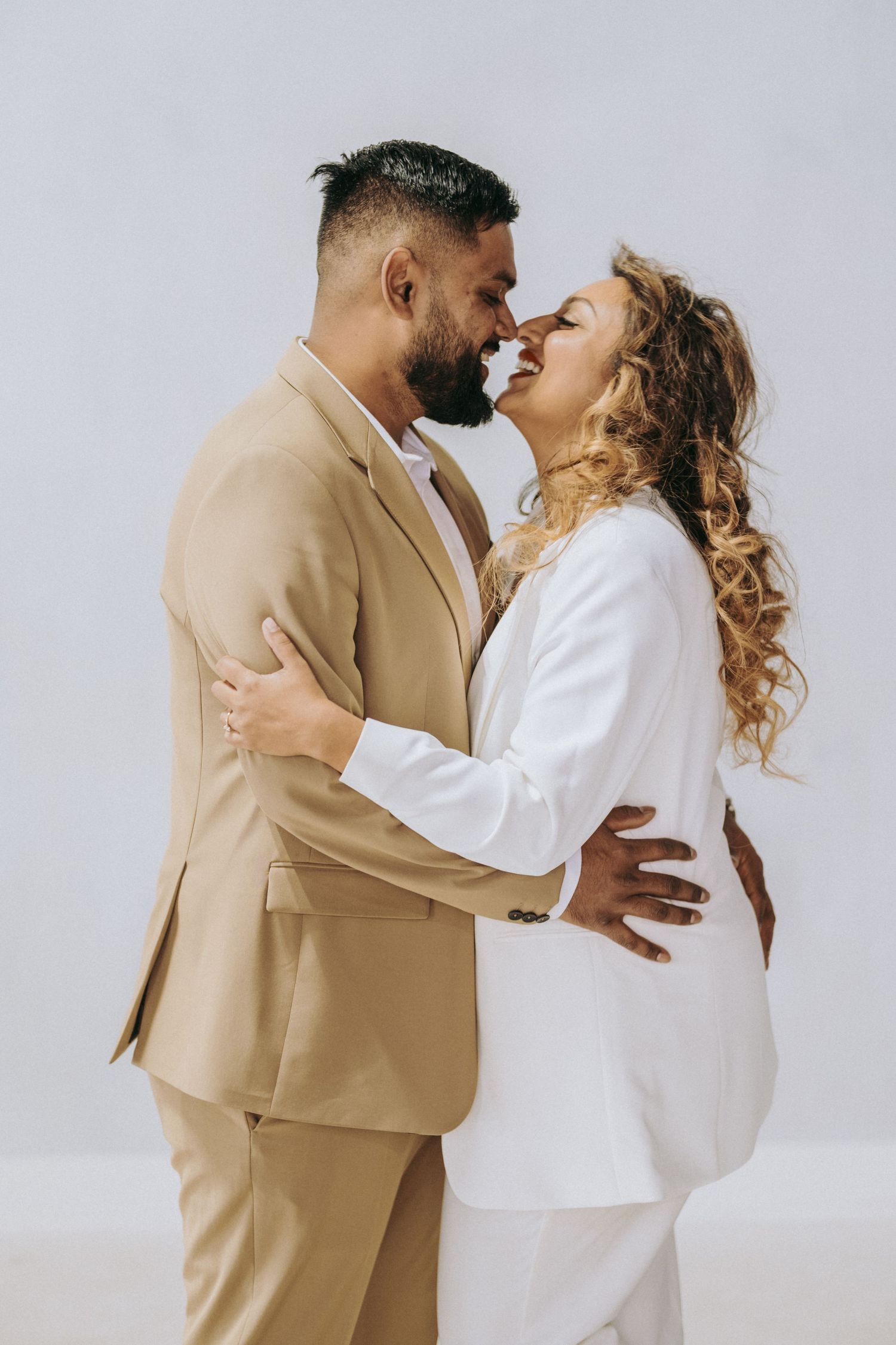 Stylish couple in beige and white suits share intimate moment against neutral background.