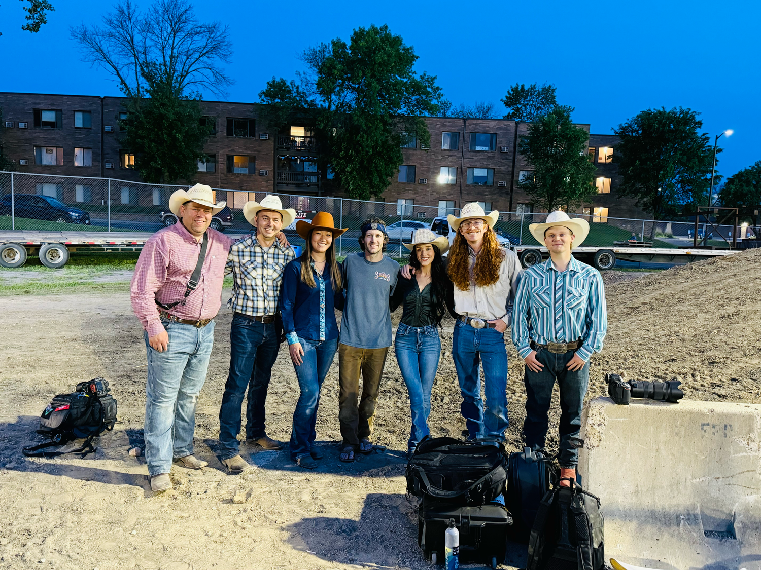 A group in western attire stands together at dusk in front of a brick building.