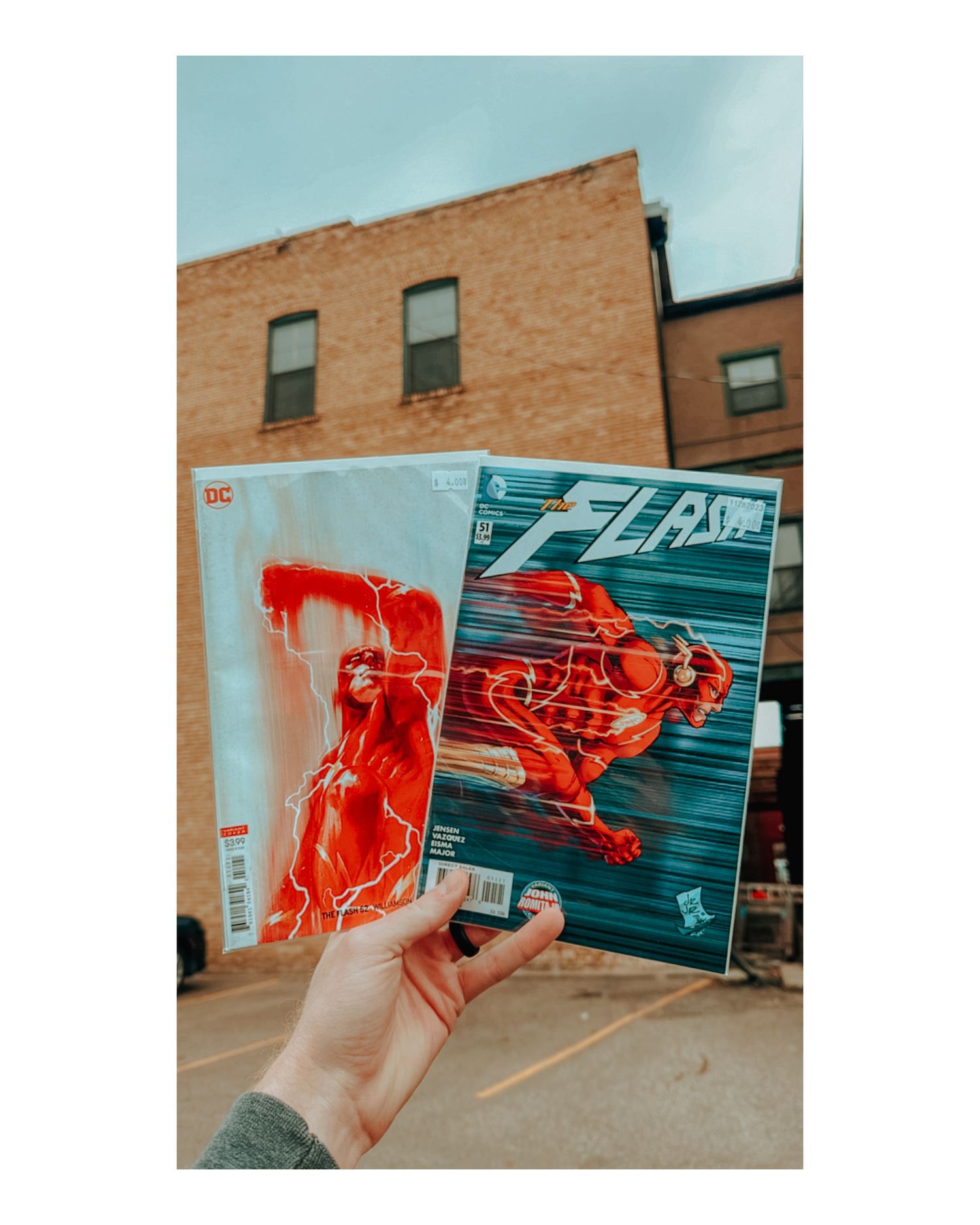 Hand holding comic books featuring red superhero artwork against brick wall background.