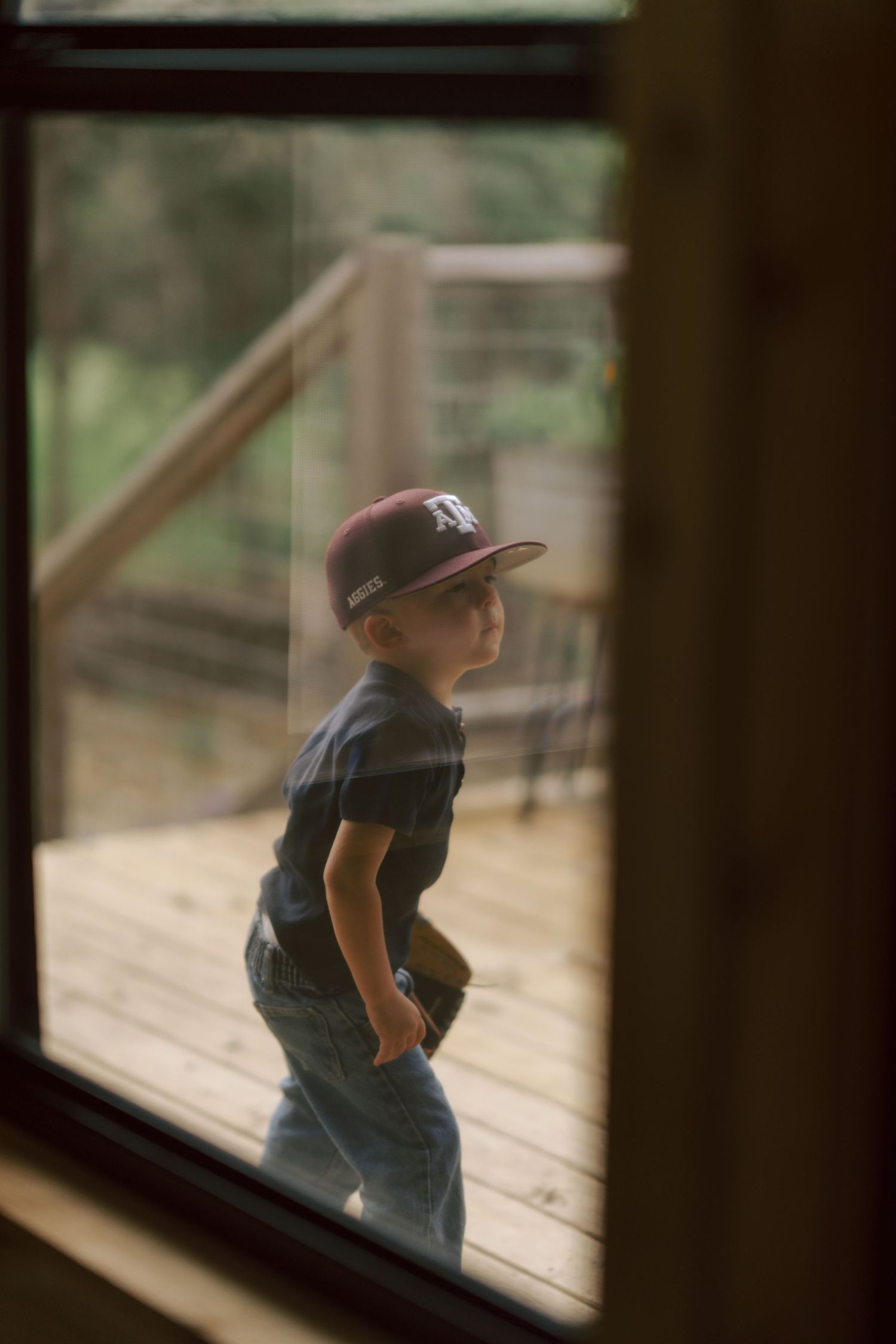 Young child wearing a baseball cap viewed through a window while standing on a wooden deck outside.