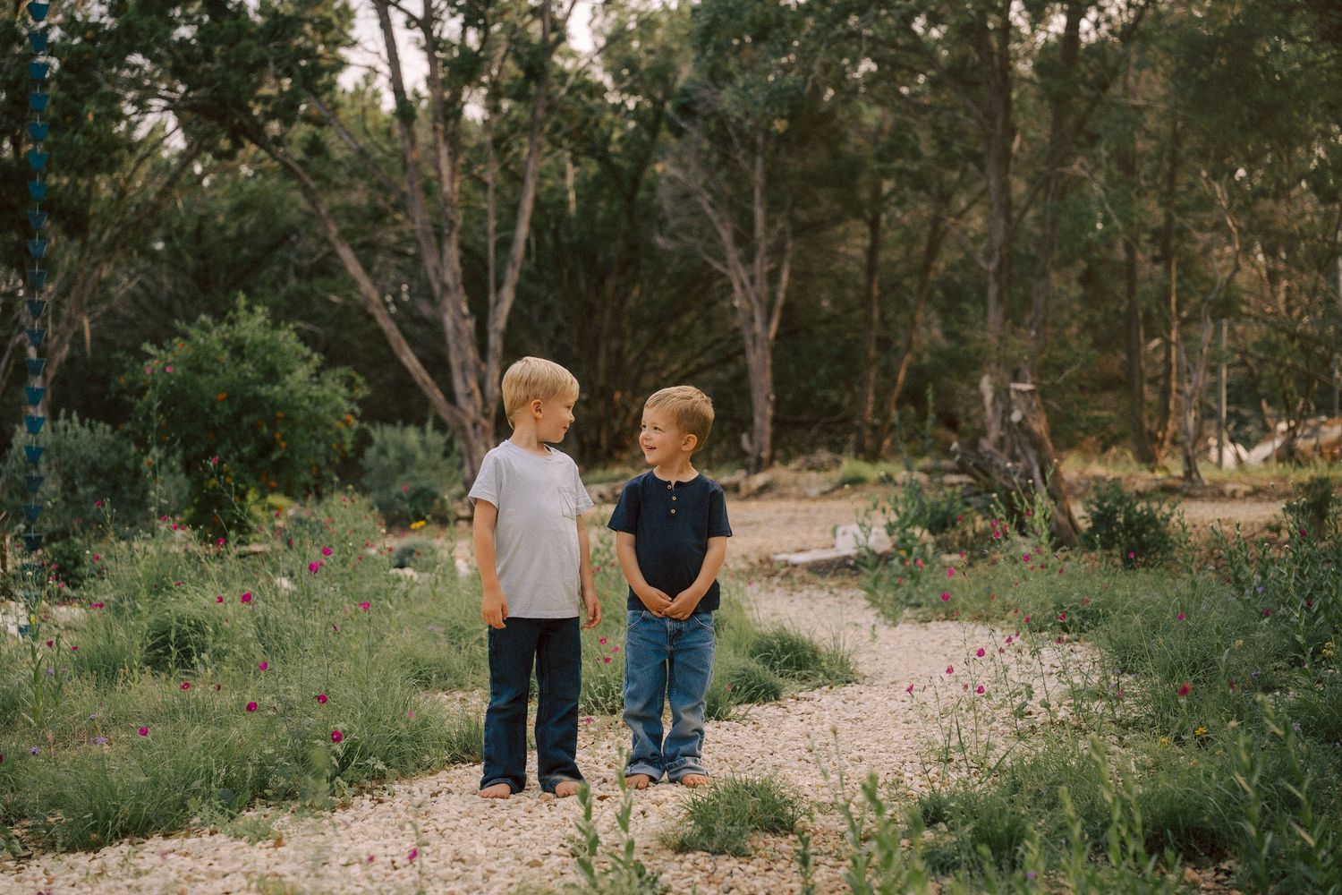 Two children standing together on a gravel path in a wooded natural setting during golden hour.