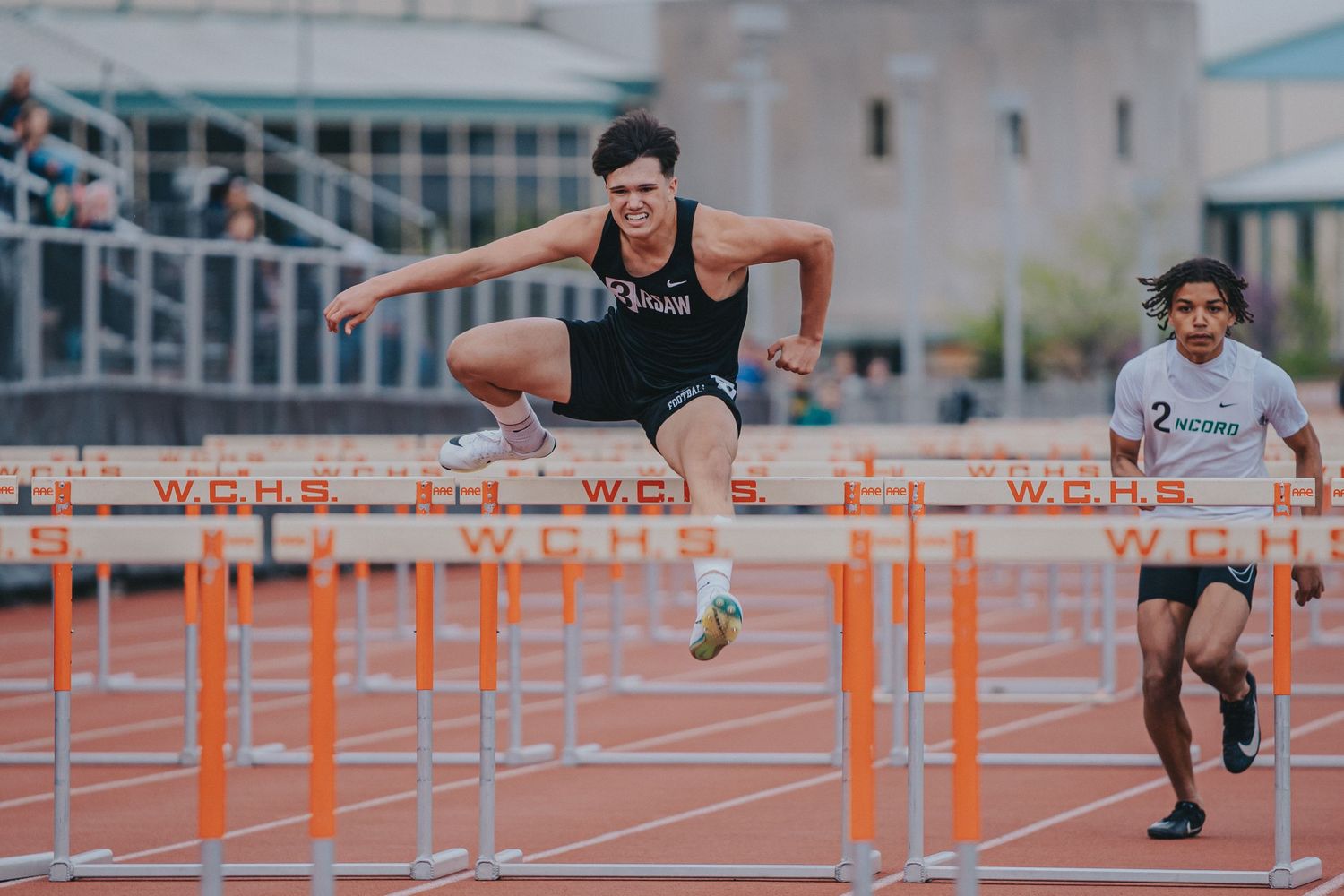 Track athlete clearing orange hurdles during competitive race at outdoor stadium.