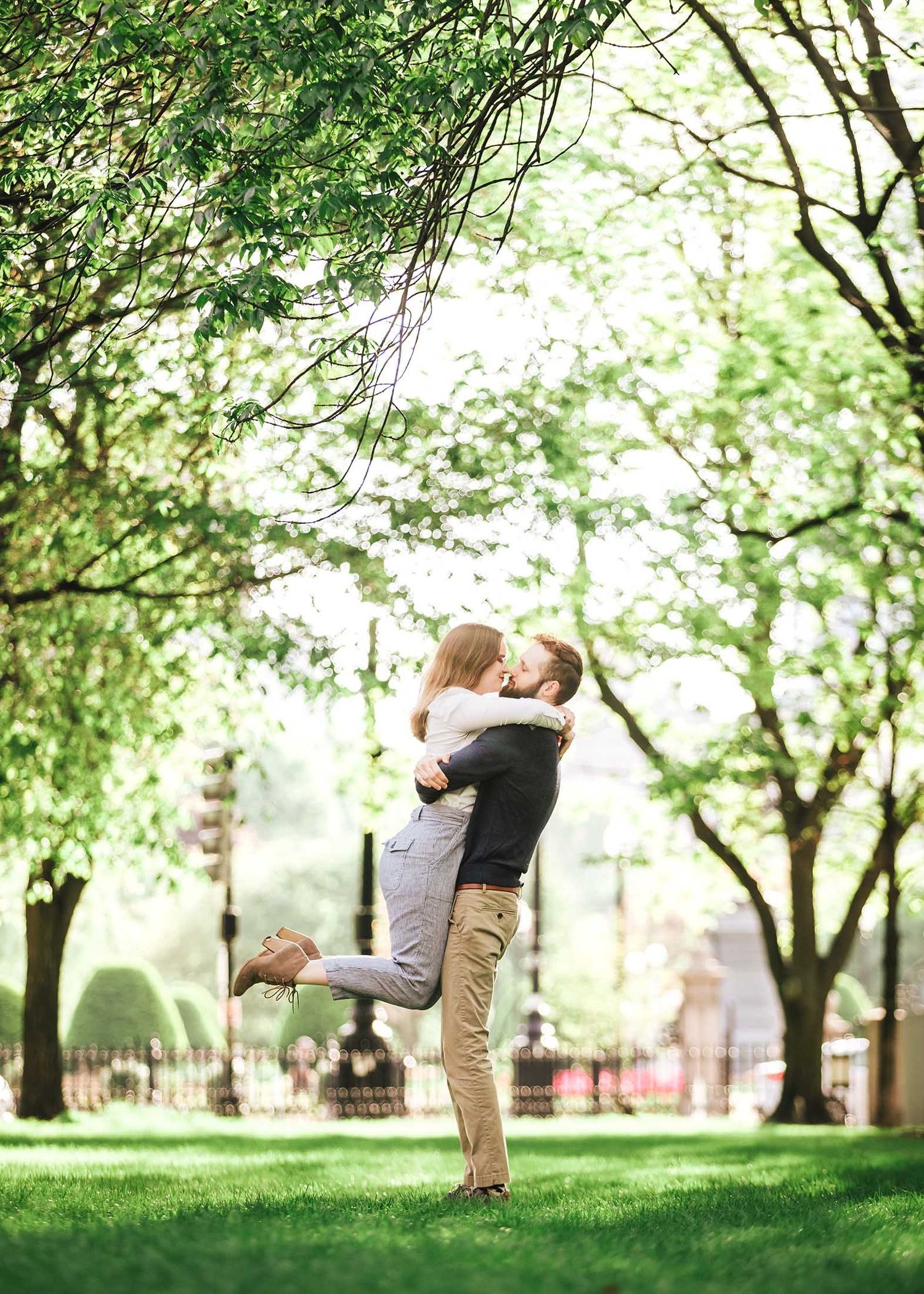 Groom-to-be lifting and embracing fiancée with joyful expression on Commonwealth Avenue Mall in Boston engagement photos