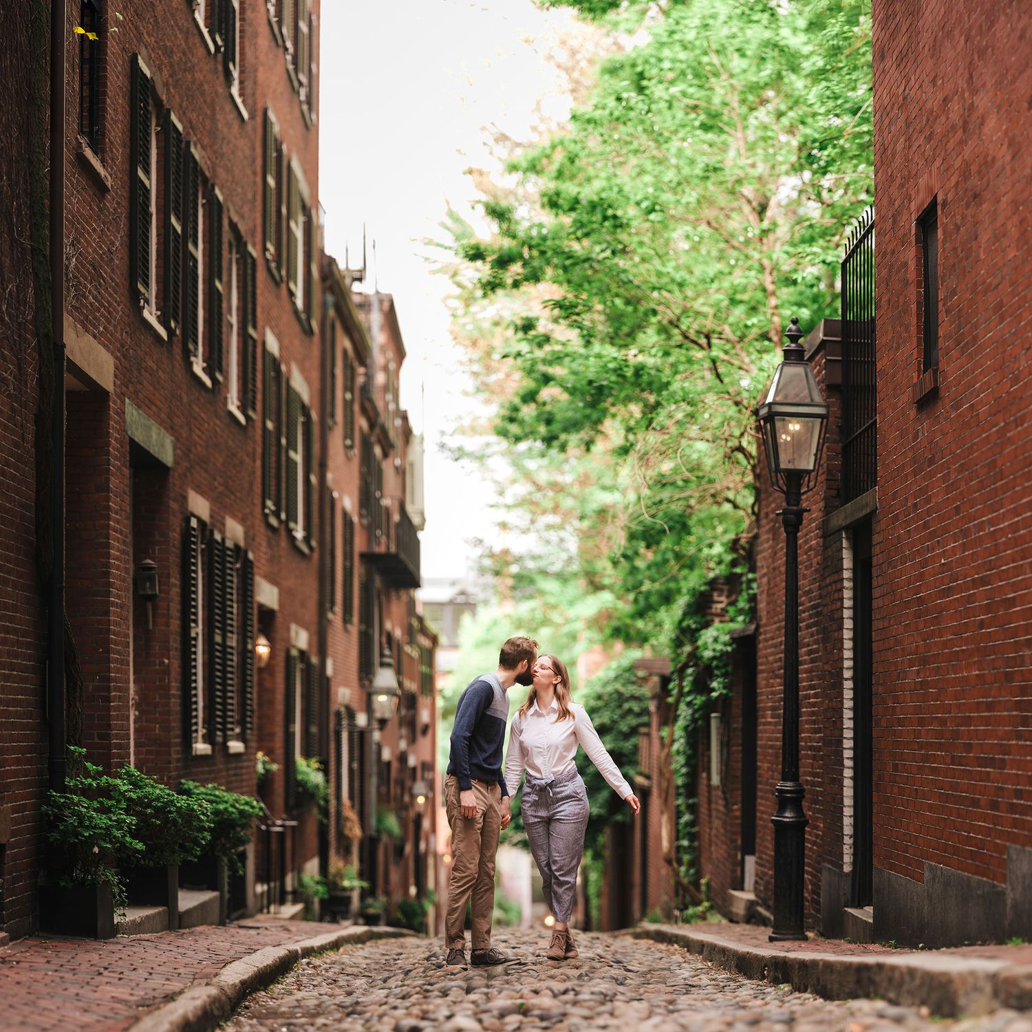A wide-angle portrait of a couple sharing a kiss in the middle of Acorn Street’s cobblestones, framed by lush green trees and red brick