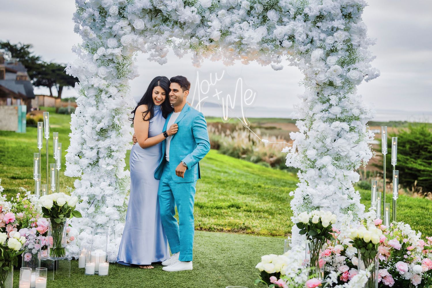 A couple poses under an elegant white floral arch at an outdoor wedding venue with candles and flowers.