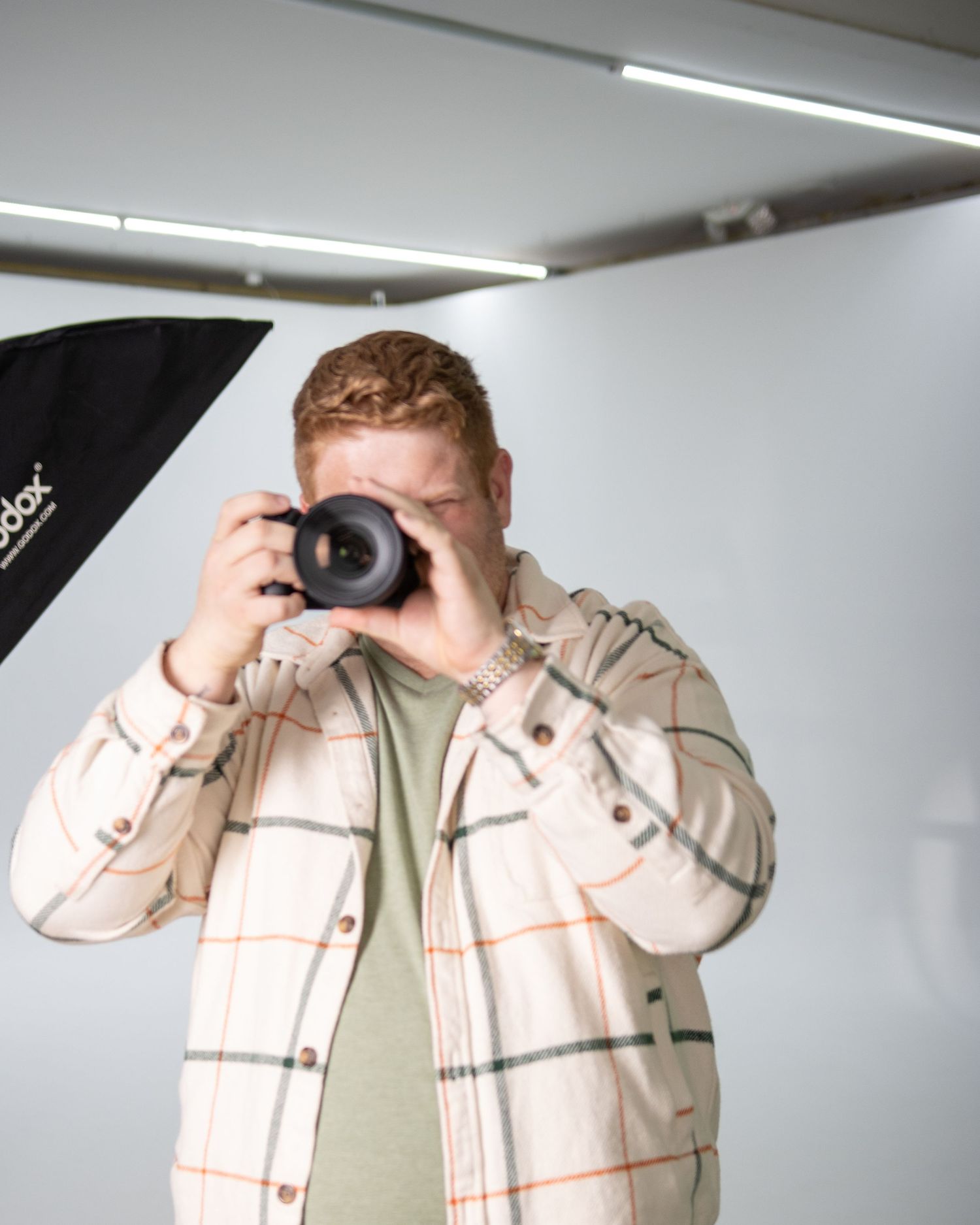 A photographer in a plaid shirt takes a photo with a professional camera in a studio setting with softbox lighting.