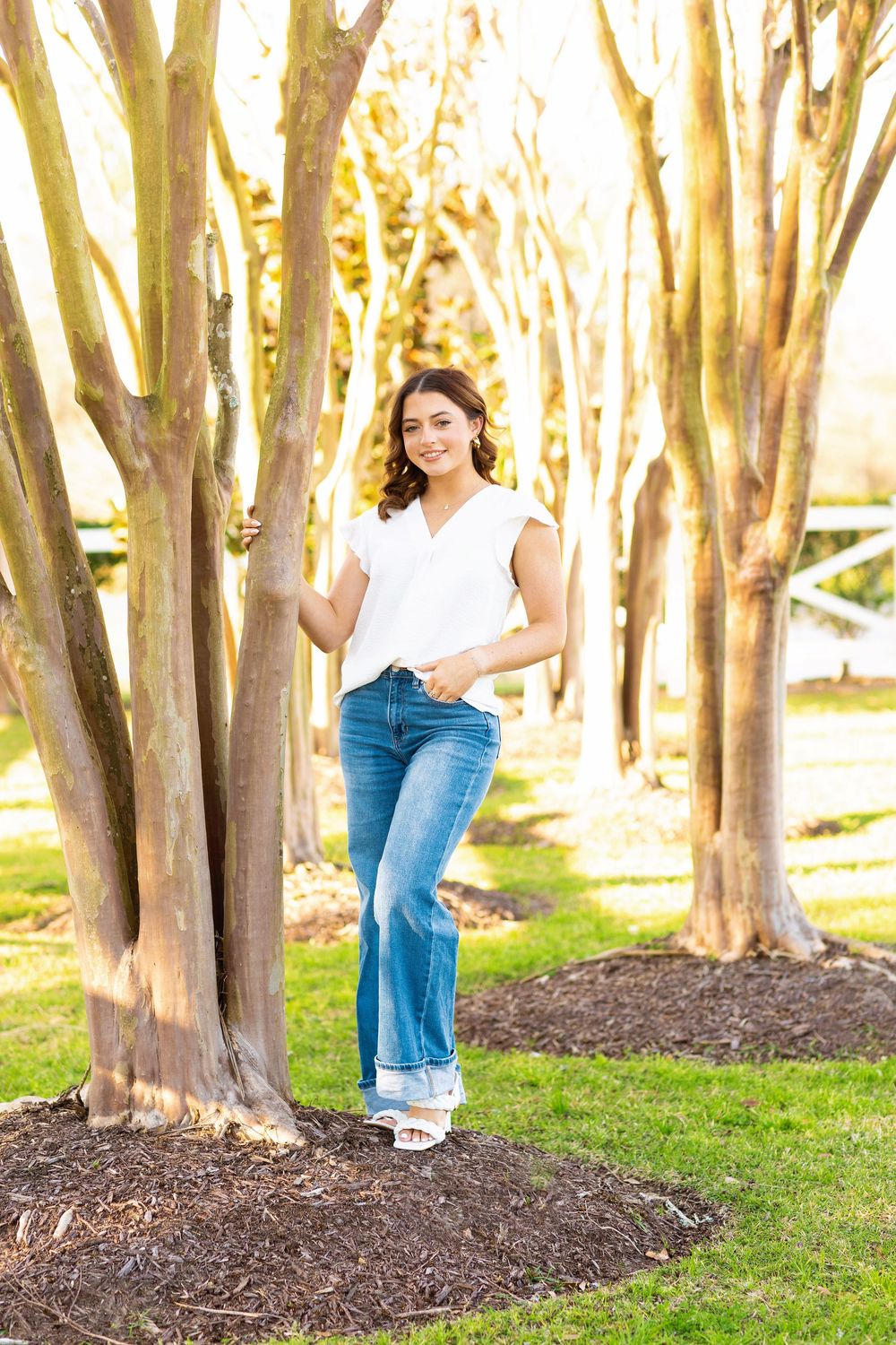 A person stands casually in a sunlit grove of crepe myrtle trees wearing a white top and blue jeans.