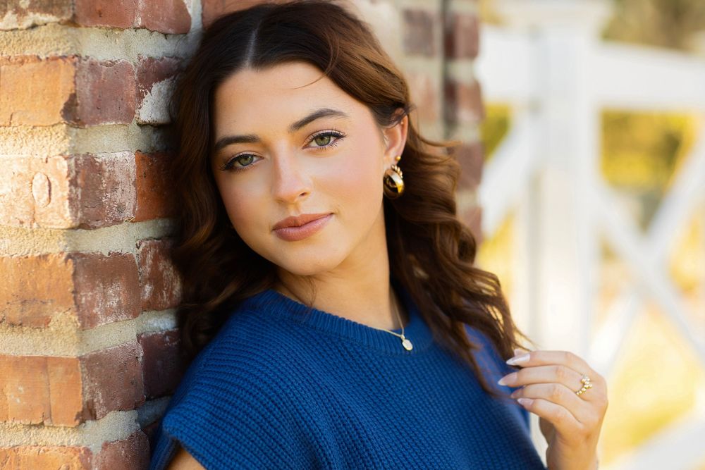 A person in a blue sweater standing against a brick wall with warm outdoor lighting and soft focus background.