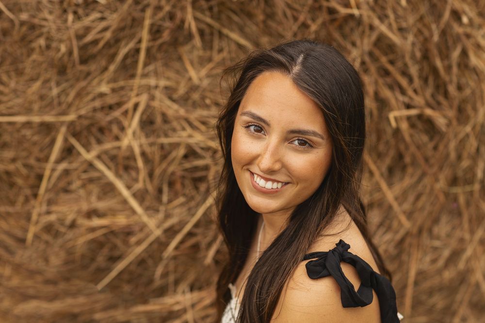 A smiling person with long dark hair poses near a hay bale wearing a sleeveless white top with black shoulder detail.