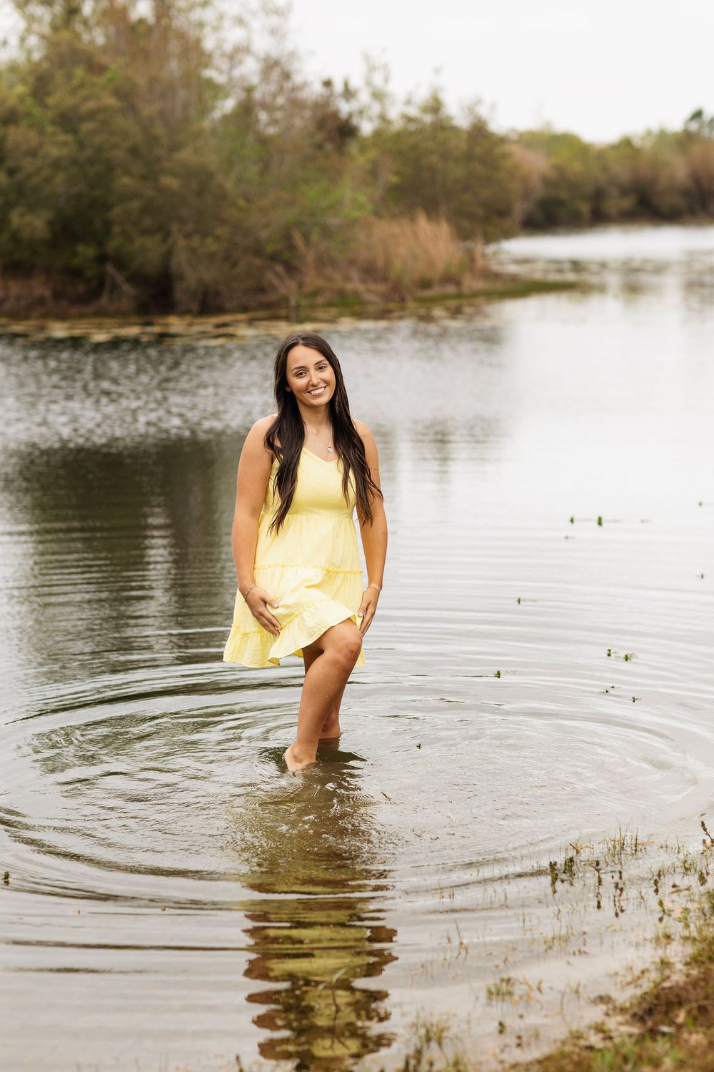 Person in yellow sundress stands knee-deep in tranquil lake waters with scenic wooded shoreline in background.