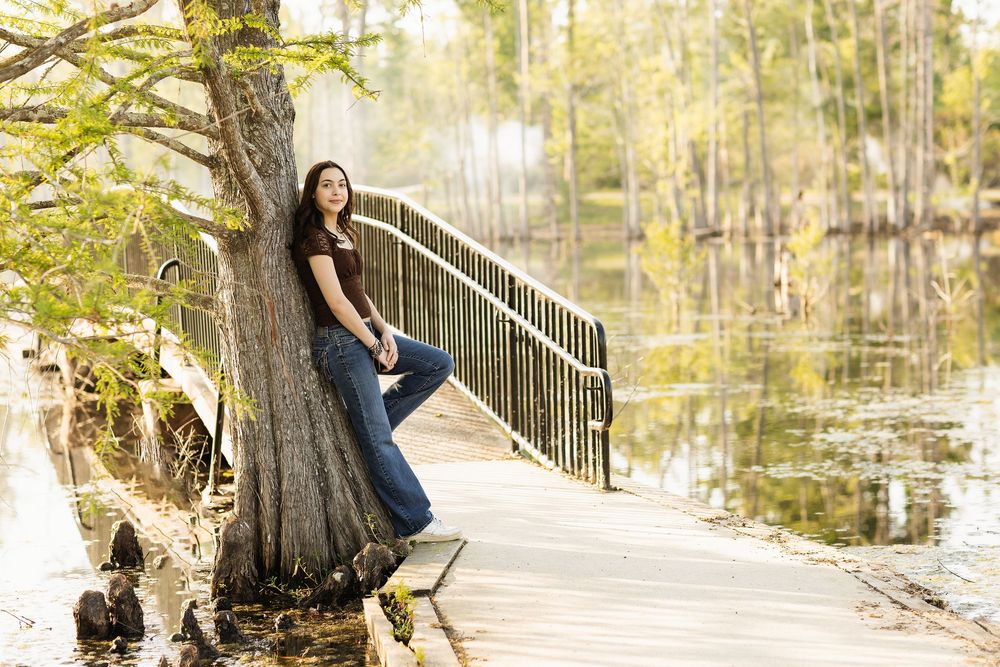 Someone in blue jeans leans against a cypress tree on a wooden boardwalk path over tranquil swamp waters in autumn.