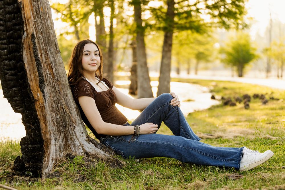Young person in casual attire relaxes against tree trunk in sunlit park setting during golden hour.
