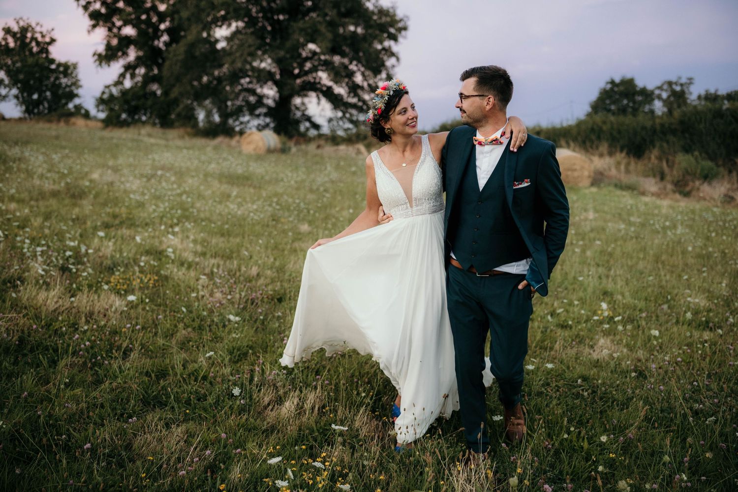 A couple in wedding attire stands together in a grassy meadow with trees in the background under moody skies.