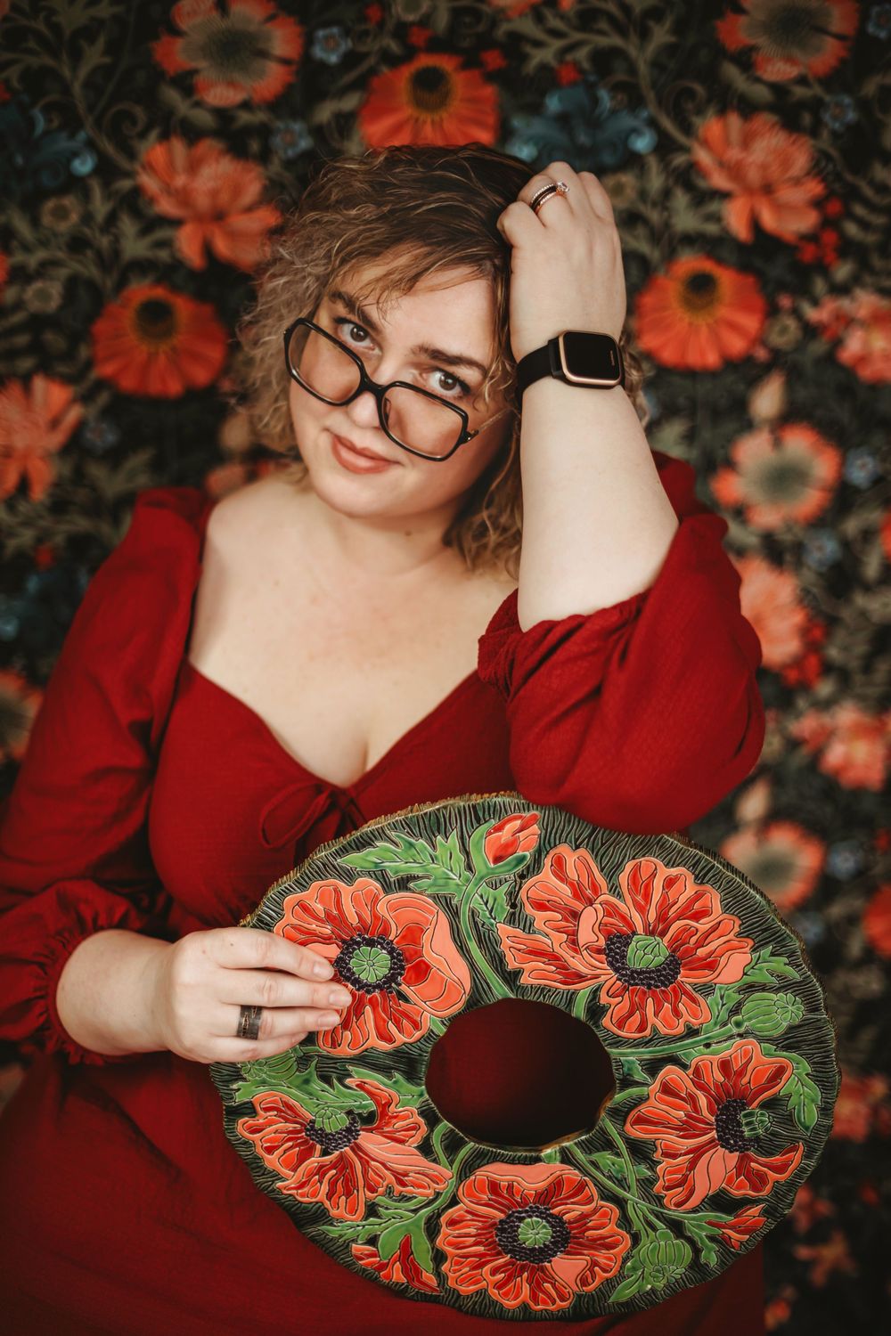Model in red dress and glasses holds decorative orange poppy wreath against floral patterned background.